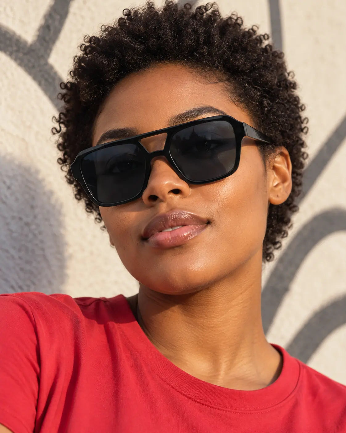 Mulher de cabelo curto usando óculos de sol feminino quadrado preto Vittoria LURVANI, com lentes fumê e ponte vazada, diante de parede urbana.