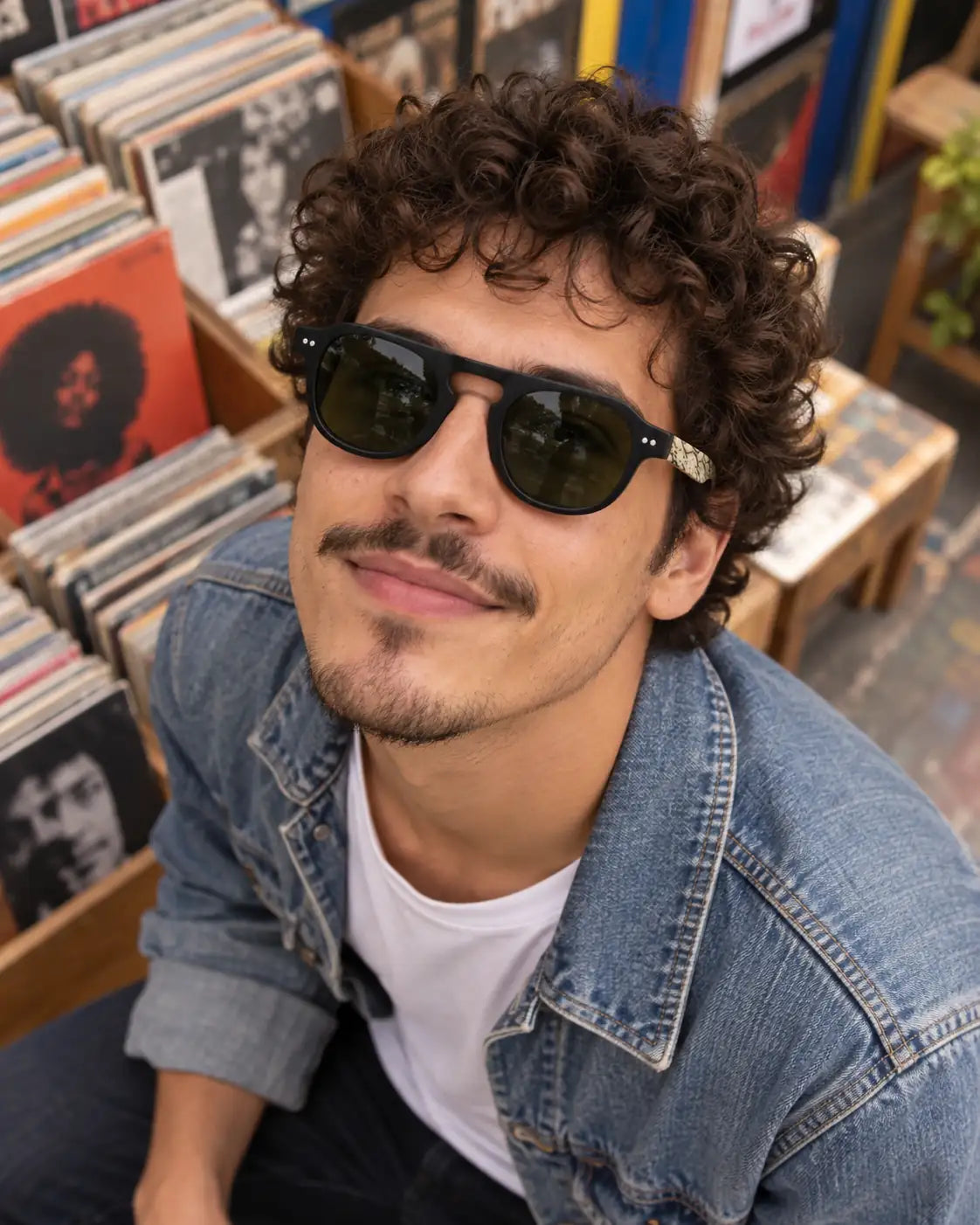 Homem de cabelos cacheados usando óculos de sol masculino quadrado preto Verona LURVANI, com lentes verdes e hastes marmorizadas, em loja de discos.